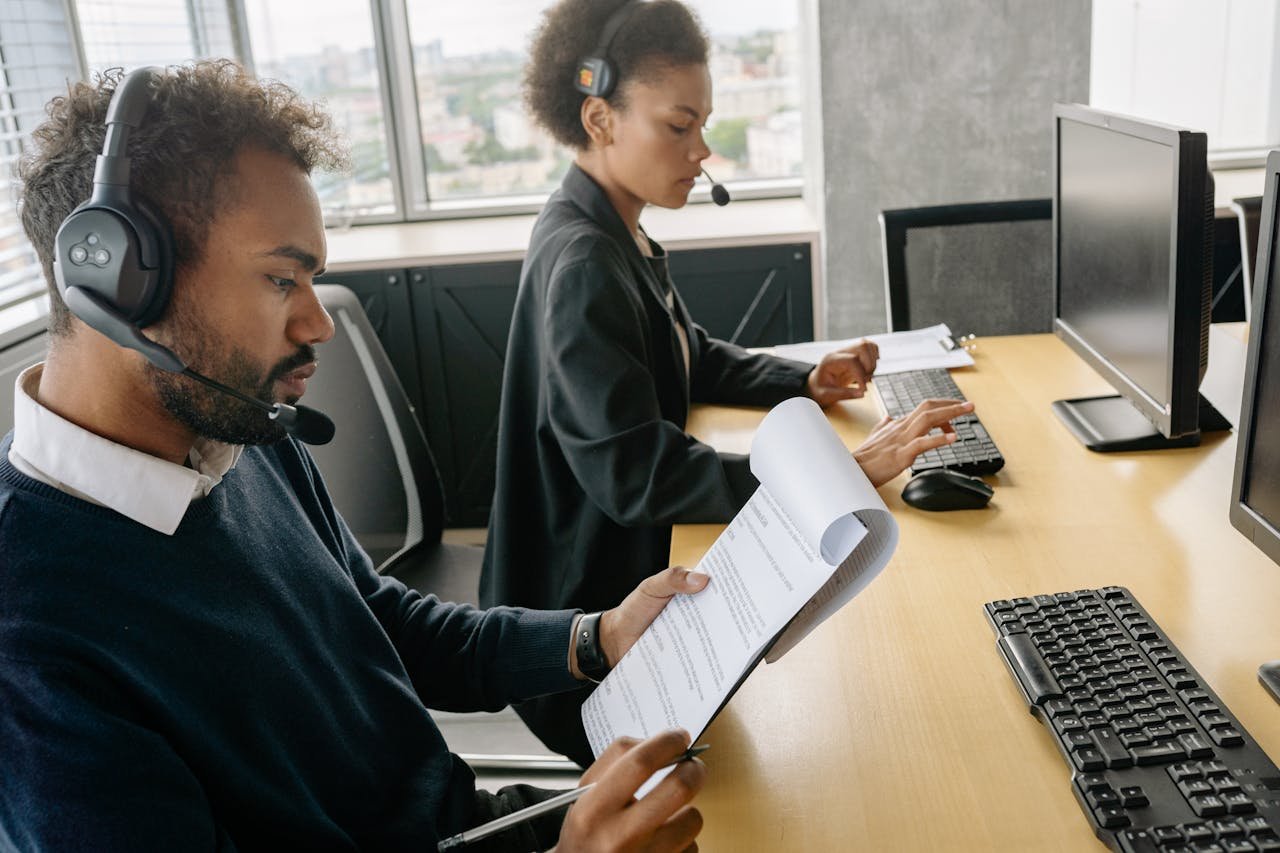 Two professionals working at a call center, wearing headsets, reading documents.
